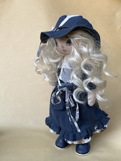 A handcrafted doll with long, curly blonde hair wearing a dark blue, wide-brimmed hat and a matching outfit. The dress is adorned with ribbons and the shoes are also blue. The doll's facial features are delicately embroidered, adding to its charming appearance.
