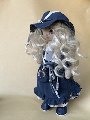 A handcrafted doll with long, curly blonde hair wearing a dark blue, wide-brimmed hat and a matching outfit. The dress is adorned with ribbons and the shoes are also blue. The doll's facial features are delicately embroidered, adding to its charming appearance.