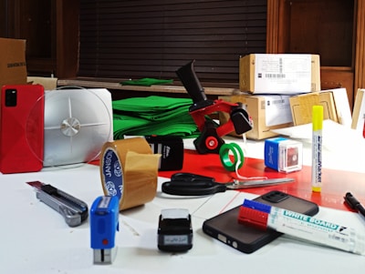 A collection of various office supplies and packages are scattered across a table. Items include a red phone, a serrated knife, rolls of packing tape, scissors, markers, a stapler, and several cardboard boxes. The background features a stack of green material, possibly paper or fabric, and a window with blinds.