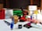 A collection of various office supplies and packages are scattered across a table. Items include a red phone, a serrated knife, rolls of packing tape, scissors, markers, a stapler, and several cardboard boxes. The background features a stack of green material, possibly paper or fabric, and a window with blinds.