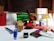 A collection of various office supplies and packages are scattered across a table. Items include a red phone, a serrated knife, rolls of packing tape, scissors, markers, a stapler, and several cardboard boxes. The background features a stack of green material, possibly paper or fabric, and a window with blinds.
