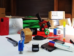 A collection of various office supplies and packages are scattered across a table. Items include a red phone, a serrated knife, rolls of packing tape, scissors, markers, a stapler, and several cardboard boxes. The background features a stack of green material, possibly paper or fabric, and a window with blinds.