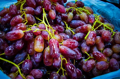 A pile of fresh, reddish-purple grapes with glossy skins and vibrant green stems gathered together, filling the frame. The grapes exhibit varied hues from deep maroon to lighter pink tones, and they appear plump and juicy.