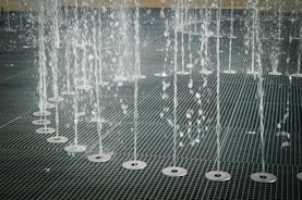 Water jets shooting upwards in an organized row across a metal grate surface, creating a pattern of streaming water droplets.