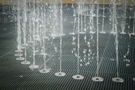 Water jets shooting upwards in an organized row across a metal grate surface, creating a pattern of streaming water droplets.