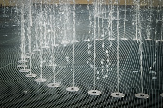Water jets shooting upwards in an organized row across a metal grate surface, creating a pattern of streaming water droplets.