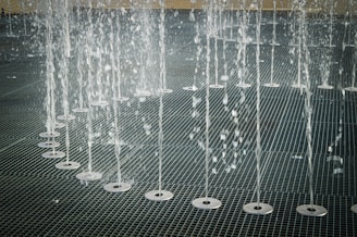 Water jets shooting upwards in an organized row across a metal grate surface, creating a pattern of streaming water droplets.