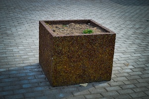 A large, textured cubic planter sits on a paved stone surface. The planter is mostly empty, containing some dirt and a small patch of green plants. The pavement consists of rectangular grey stones laid in a uniform pattern.