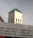 A church tower with a cross on top is visible above a building with a blue roof. In the foreground, there is a partially visible sign featuring text advertising various money transfer services and printing-related activities.