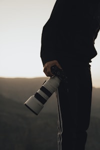 A person holding a large camera with a telephoto lens, framed against a backdrop of hazy mountains. The camera is held in one hand, pointing downward, with the person dressed in dark clothing, including a jacket with white stripes on the pants.