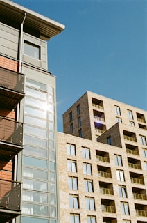 Modern urban architecture featuring two multi-story buildings with numerous glass windows and balconies. One building has a sleek grey exterior with reflections of sunlight on the glass, while the other has a brick facade.