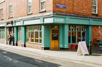 A brick building with teal-colored shopfront, featuring large windows and a wooden door with a sign reading 'SIOP = SHOP'. The shop overlooks a quiet street with a sidewalk. A sandwich board with 'SIOP' written on it stands outside. To the side, a woman is talking to a young child. Graffiti is visible on the wall, along with a blue street sign reading 'SILVER JUBILEE WALK'.