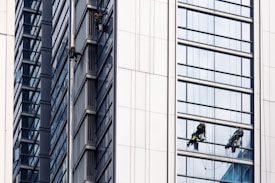 Several workers are suspended on the side of a modern skyscraper, engaged in cleaning the windows. They are equipped with safety harnesses and ropes, operating from different heights along the building's facade. The building's surface features reflective glass panels that capture the surrounding environment.
