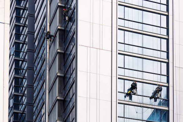 Several workers are suspended on the side of a modern skyscraper, engaged in cleaning the windows. They are equipped with safety harnesses and ropes, operating from different heights along the building's facade. The building's surface features reflective glass panels that capture the surrounding environment.