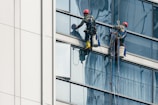 Two workers wearing safety gear are suspended by ropes on the side of a glass building, cleaning the windows. They are equipped with buckets and tools as they perform their task at significant heights.