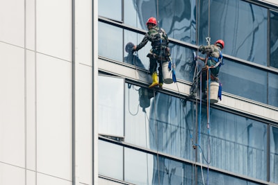 Two workers wearing safety gear are suspended by ropes on the side of a glass building, cleaning the windows. They are equipped with buckets and tools as they perform their task at significant heights.