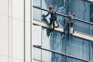 Two workers wearing safety gear are suspended by ropes on the side of a glass building, cleaning the windows. They are equipped with buckets and tools as they perform their task at significant heights.