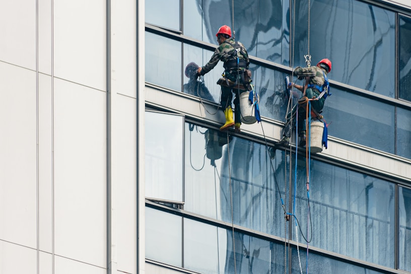 Two workers wearing safety gear are suspended by ropes on the side of a glass building, cleaning the windows. They are equipped with buckets and tools as they perform their task at significant heights.