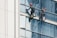 Two workers wearing safety gear are suspended by ropes on the side of a glass building, cleaning the windows. They are equipped with buckets and tools as they perform their task at significant heights.