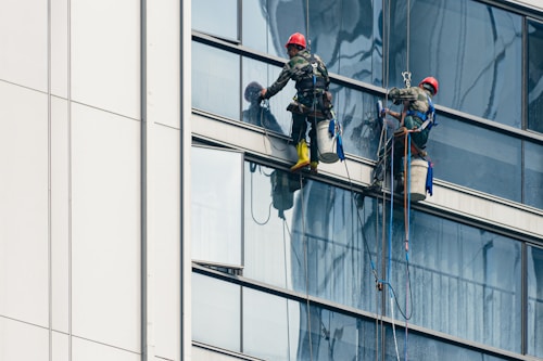 Two workers wearing safety gear are suspended by ropes on the side of a glass building, cleaning the windows. They are equipped with buckets and tools as they perform their task at significant heights.