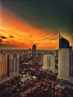 A vibrant cityscape during sunset with a striking mix of high-rise buildings and a moody sky. The skyline features modern architecture with distinctive skyscrapers set against a dramatic, richly colored sky transitioning from golden orange to deep shadows.