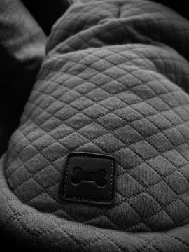 A close-up of a quilted fabric surface featuring a small, square patch with a bone symbol. The texture of the quilted pattern is prominent, highlighting the stitching and fabric folds. The image predominantly uses a black and white color scheme.