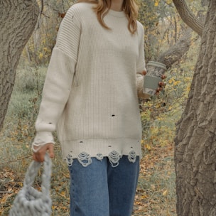 A person stands in an outdoor setting surrounded by trees and autumn foliage, wearing a cream-colored, distressed sweater and blue jeans. They hold a textured, reusable coffee cup and a knitted bag, suggesting a casual, relaxed mood.
