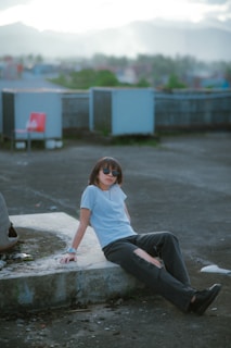 A person wearing sunglasses and casual clothing is sitting on a concrete surface outdoors. The setting appears to be an urban rooftop with mountains visible in the background under a cloudy sky. The person is leaning back with one arm resting and looking relaxed.