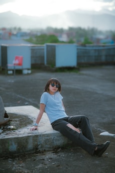 A person wearing sunglasses and casual clothing is sitting on a concrete surface outdoors. The setting appears to be an urban rooftop with mountains visible in the background under a cloudy sky. The person is leaning back with one arm resting and looking relaxed.