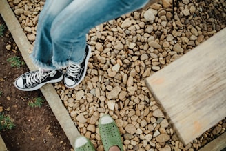 A close-up view of a pair of black sneakers with white soles and a pair of green shoes with perforated holes. The feet are on a gravel walkway bordered by a strip of soil with small patches of grass. A wooden bench is partially visible in the top right corner.