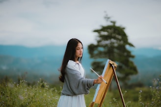 A person is painting on an easel outdoors in a natural setting. The background shows a blurry landscape with mountains and a large tree, giving a serene and picturesque scene.
