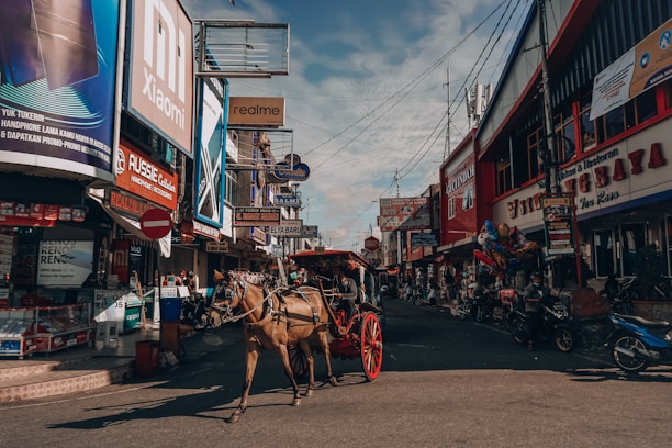 A bustling city street filled with various storefronts and large signs advertising different brands, including Xiaomi and realme. A horse-drawn carriage is prominently featured in the middle of the street, with the horse and cart casting shadows on the pavement. Numerous motorcycles and people can be seen on the street, adding to the lively urban atmosphere.