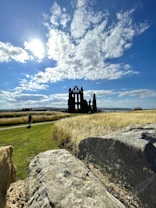 The image depicts a scenic view of an ancient abbey silhouetted against a bright sky. Puffy white clouds scatter across the blue expanse, while the sun shines brilliantly, casting its light onto the landscape. In the foreground, large stones and a path are surrounded by tall golden grass that sways gently in the open field. A lone figure can be seen walking on the path, adding a sense of scale to the vast surroundings.