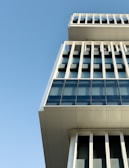 A modern building with a unique architectural design, featuring large glass windows and a series of vertical and horizontal lines. The structure juts out at the top, creating an interesting geometric perspective against a clear blue sky.