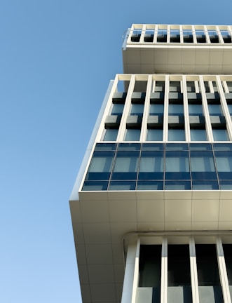 A modern building with a unique architectural design, featuring large glass windows and a series of vertical and horizontal lines. The structure juts out at the top, creating an interesting geometric perspective against a clear blue sky.