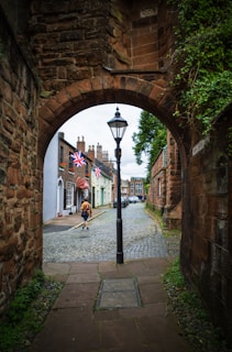 An archway leads to a cobblestone street lined with charming buildings. A streetlamp stands prominently in the center, and a person wearing an orange jacket walks down the street. British flags hang from the buildings, and there’s greenery climbing along the stone walls.