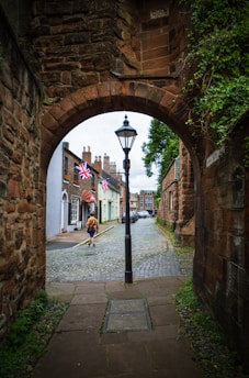 An archway leads to a cobblestone street lined with charming buildings. A streetlamp stands prominently in the center, and a person wearing an orange jacket walks down the street. British flags hang from the buildings, and there’s greenery climbing along the stone walls.