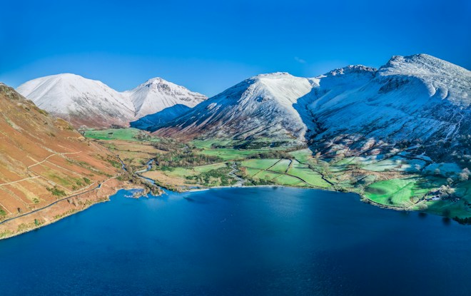 Snow-capped mountains rise majestically above a tranquil blue lake, surrounded by lush green fields and winding roads. The contrasting colors emphasize the vibrant beauty of the landscape.