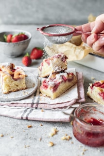 A hand sprinkles powdered sugar over slices of cake topped with fresh strawberries, elegantly arranged on a plate with a red-striped cloth beneath. A bowl of fresh strawberries is in the background, adding a vibrant touch next to the glass jar filled with strawberry jam visible at the bottom right.