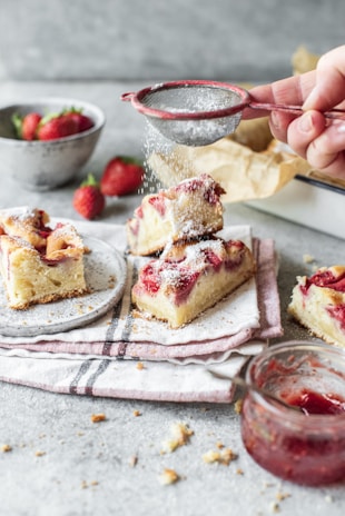 A hand sprinkles powdered sugar over slices of cake topped with fresh strawberries, elegantly arranged on a plate with a red-striped cloth beneath. A bowl of fresh strawberries is in the background, adding a vibrant touch next to the glass jar filled with strawberry jam visible at the bottom right.