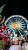 A vibrant carnival scene at night with a large, illuminated Ferris wheel spinning rapidly in the background. The foreground features colorful stalls and vibrant decorations, including bright lights and ornate structures. People are walking along the pathway, adding a lively atmosphere to the scene.