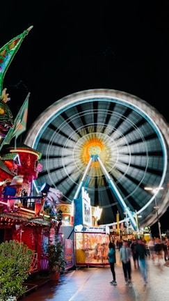 A vibrant carnival scene at night with a large, illuminated Ferris wheel spinning rapidly in the background. The foreground features colorful stalls and vibrant decorations, including bright lights and ornate structures. People are walking along the pathway, adding a lively atmosphere to the scene.
