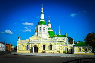 A beautiful church with distinctive green and white domes set against a bright blue sky. The building features elegant architectural details, including arched windows and golden crosses on top of the domes. Surrounding the structure is a modest urban environment, with a few trees and a road in the foreground.
