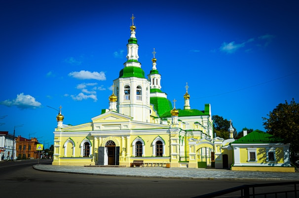 A beautiful church with distinctive green and white domes set against a bright blue sky. The building features elegant architectural details, including arched windows and golden crosses on top of the domes. Surrounding the structure is a modest urban environment, with a few trees and a road in the foreground.