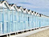 A row of identical beach huts with a light blue and white color scheme stretches along a sandy beach. The structures feature gabled roofs and are elevated on stilts. The sky is partly cloudy, adding to the serene coastal atmosphere.