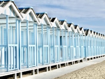 A row of identical beach huts with a light blue and white color scheme stretches along a sandy beach. The structures feature gabled roofs and are elevated on stilts. The sky is partly cloudy, adding to the serene coastal atmosphere.