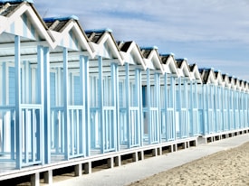 A row of identical beach huts with a light blue and white color scheme stretches along a sandy beach. The structures feature gabled roofs and are elevated on stilts. The sky is partly cloudy, adding to the serene coastal atmosphere.