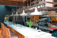 A modern bar setting featuring a long marble countertop with several wooden bar stools neatly arranged in a row. Hanging above the bar are multiple industrial-style pendant lights and an extensive collection of wine bottles and glasses organized on a rack. The bar area has various elements including a coffee machine, stacked pizza boxes, fresh plants, and decorative vases. The ambiance appears both cozy and elegant with the combination of natural wood, metal, and marble surfaces.