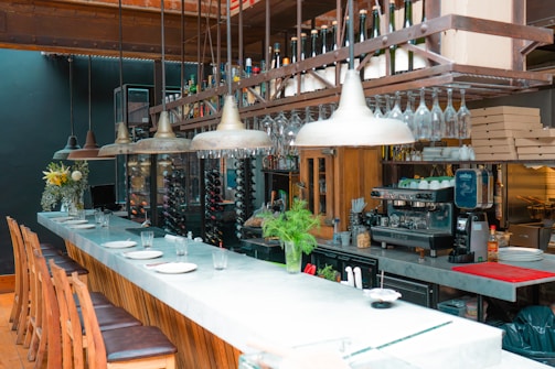 A modern bar setting featuring a long marble countertop with several wooden bar stools neatly arranged in a row. Hanging above the bar are multiple industrial-style pendant lights and an extensive collection of wine bottles and glasses organized on a rack. The bar area has various elements including a coffee machine, stacked pizza boxes, fresh plants, and decorative vases. The ambiance appears both cozy and elegant with the combination of natural wood, metal, and marble surfaces.