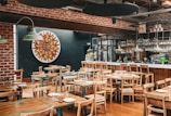 A rustic restaurant interior with wooden tables and chairs, and a brick wall. The dining area has table settings with plates and cutlery, and a bar area with various bottles and hanging wine glasses. On the wall, there's a colorful mosaic or painting of a sun face. The lighting is provided by green hanging lamps.